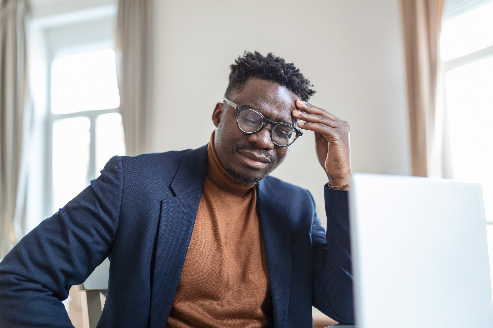 A person rubs their forehead as they view a laptop.