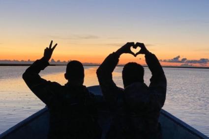 A person making heart shape and another person making a two fingers while sitting on a boat during sunset